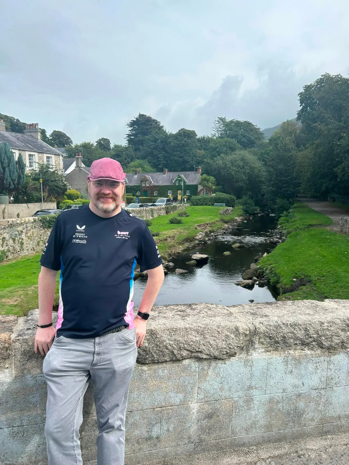 Martin Murtagh in a flat cap leaning on a stone bridge above a river, with cottages and trees behind